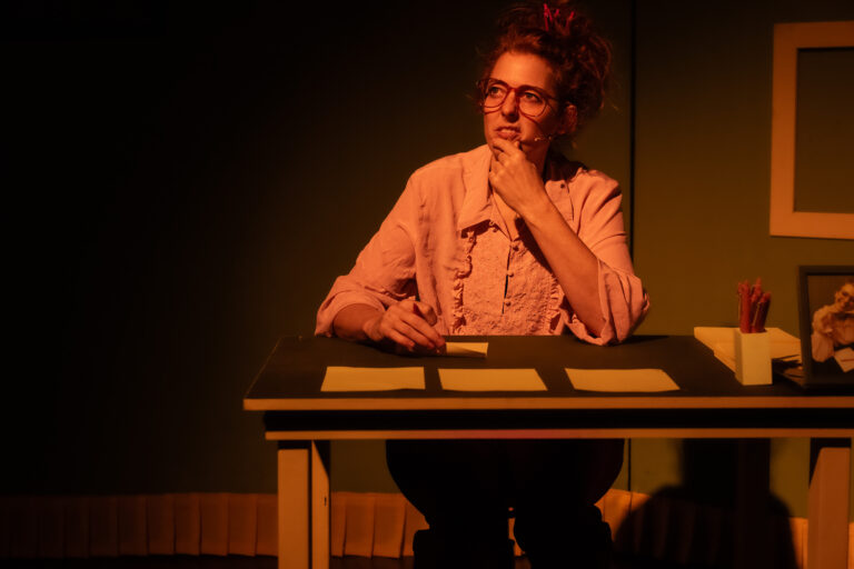 Een vrouw zit achter een bureau met papieren, terwijl ze nadenkend naar voren kijkt in een sfeervol verlichte ruimte.