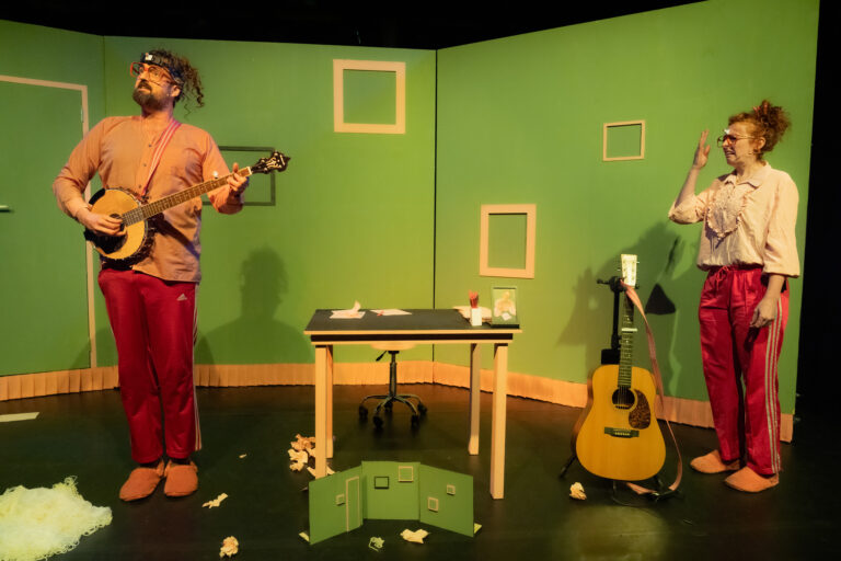 Twee performers op een kleurrige set, waarbij de man op een banjo speelt en de vrouw kijkt terwijl ze in pyjama's en slippers zijn.