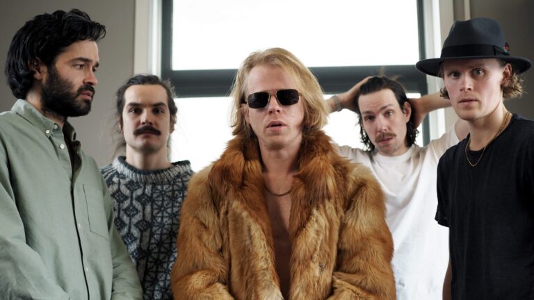 Vijf bandleden staan naast elkaar in een industriele omgeving en kijken in de camera