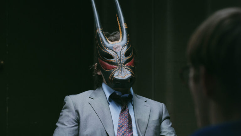 Een persoon in een grijs pak en stropdas zit aan een tafel, met een opvallend masker met lange hoorns op, terwijl een andere persoon onscherp op de voorgrond zit.