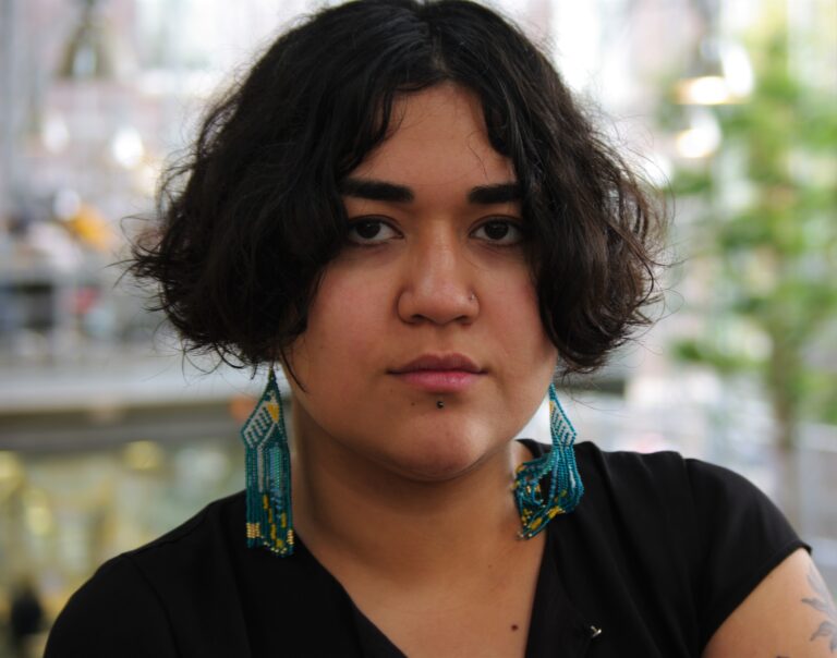 Een jonge vrouw met kort zwart haar, grote turquoise oorbellen en een zwarte top kijkt recht vooruit tegen een wazige buitenachtergrond.