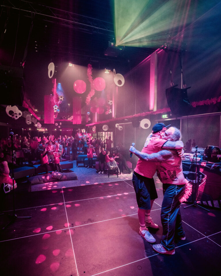 Twee mensen omarmen elkaar op een levendig dansfeest met kleurrijke verlichting en een enthousiaste menigte op de achtergrond.