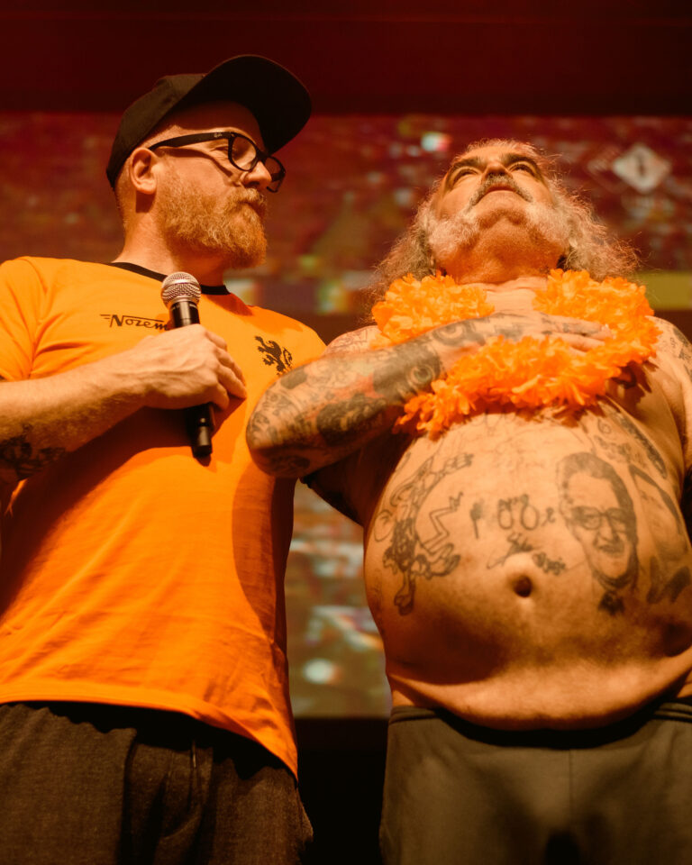 Twee mannen op een podium, één in een oranje shirt met een microfoon en de ander met een geverfd gezicht en een feloranje bloemenketting.