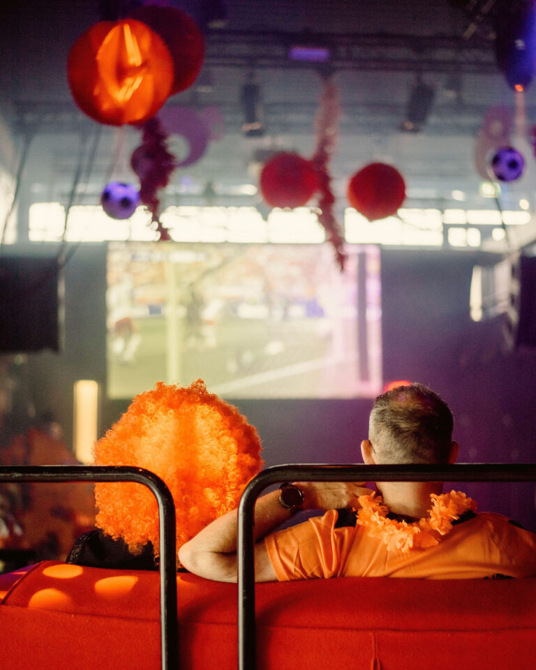 Een persoon in een oranje shirt kijkt naar een groot scherm in een feestelijke omgeving versierd met oranje decoraties.
