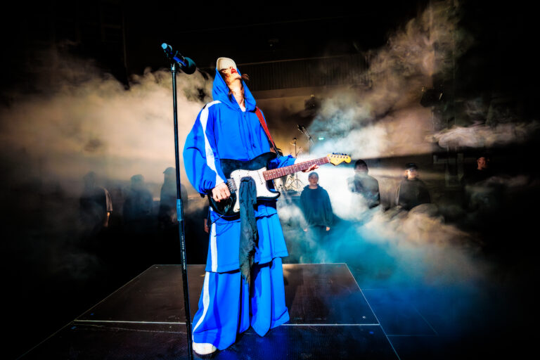 Een muzikant in een blauwe outfit speelt gitaar op een podium omringd door rook.