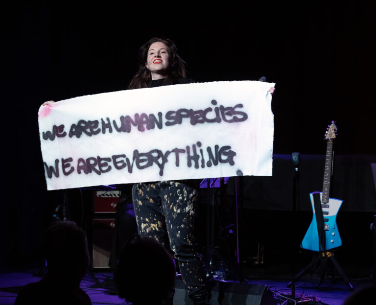 Een performer op een podium houdt een spandoek vast met de tekst