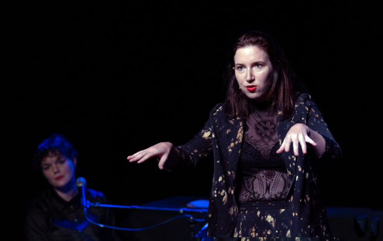 Een performer geeft een uitdrukkingrijke presentatie op een podium, met een andere persoon op de achtergrond.
