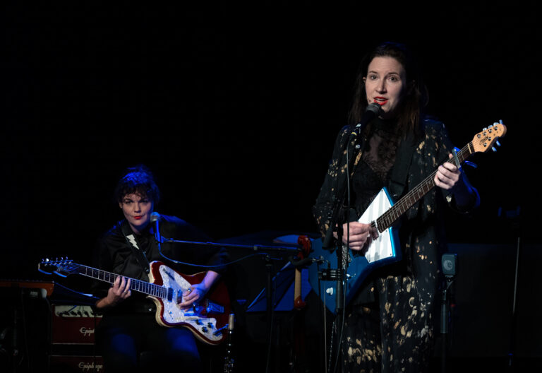 Een muzikante die een elektrische gitaar speelt en zingt, met een bassist op de achtergrond.