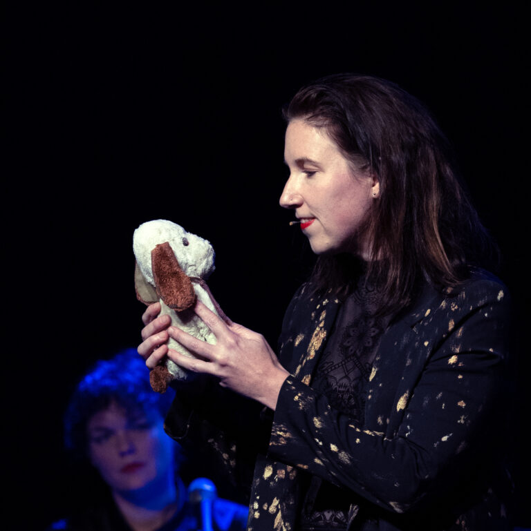 Een persoon met lang haar houdt een knuffelhondje vast op een podium, terwijl een andere persoon op de achtergrond kijkt.