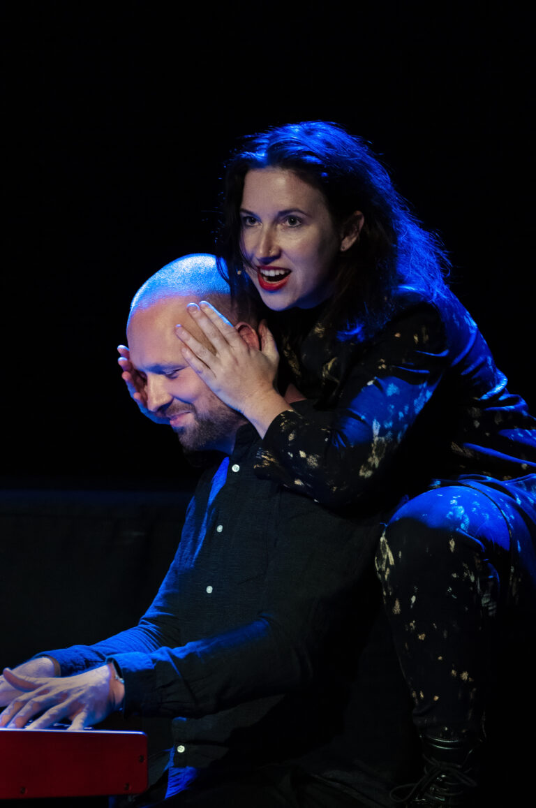 Een vrouw met donkere, golvende haren zit op de schouders van een man terwijl hij piano speelt, beiden hebben een expressieve uitstraling op het podium.