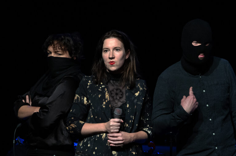 Drie performers staan op het podium, met de middelste vrouw die een microfoon vasthoudt, terwijl de anderen gedeeltelijk vermomd zijn.