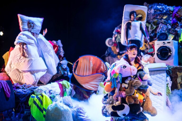 Een groep mensen rondom een kleurrijke kunstinstallatie van speelgoedachtige sculpturen op een donker podium.