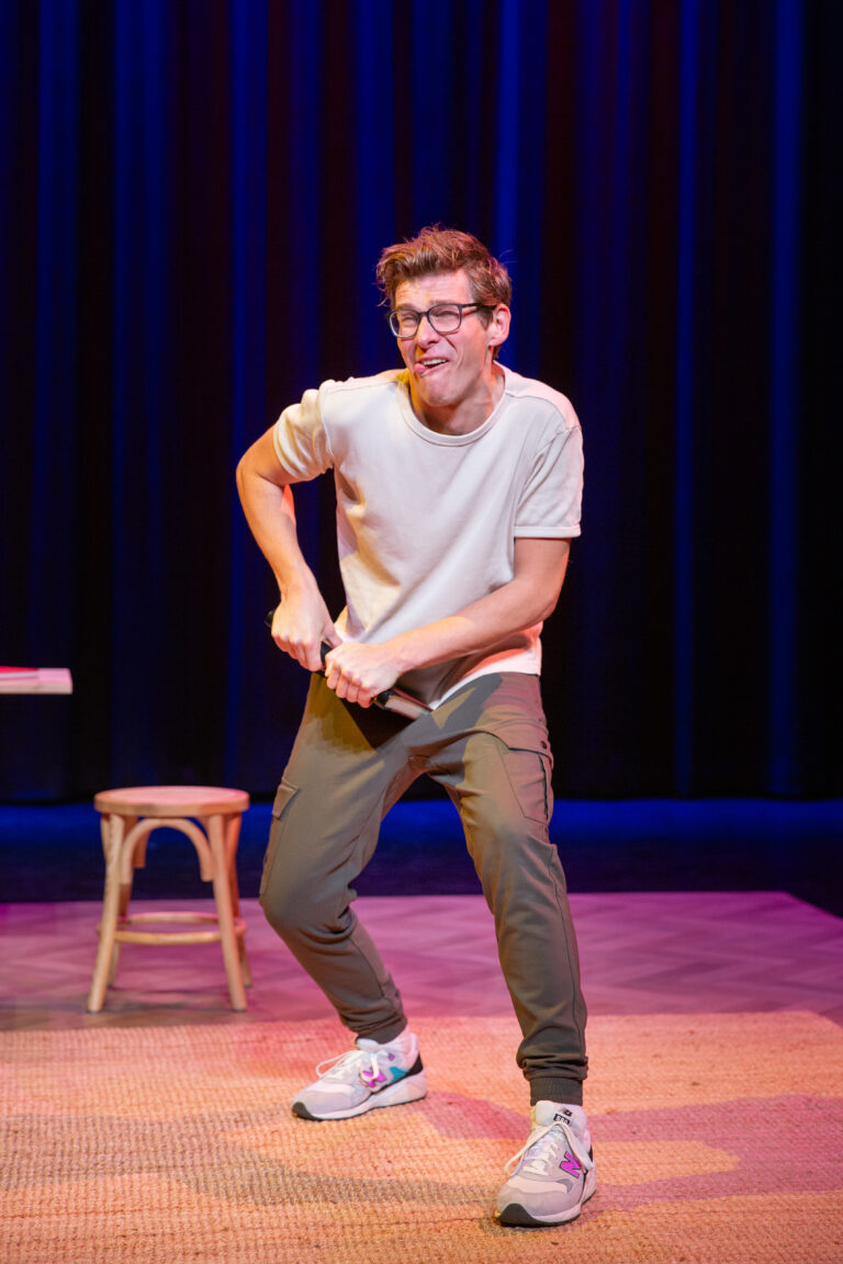 Remy staat met een wit t-shirt en een groene broek met een boek in zijn hand en tong uit zijn mond