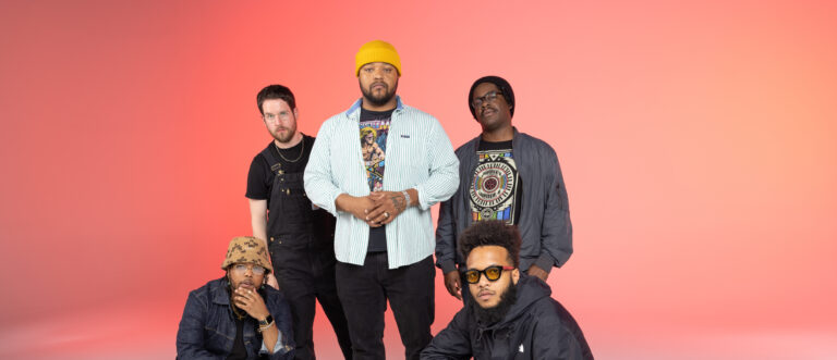 Vijf mannen poseren samen voor een roze achtergrond; drie van hen staan rechtop en twee hurken, met een gevarieerde mix van stijlen en accessoires zoals een gele muts en een hoed met camouflageprint.