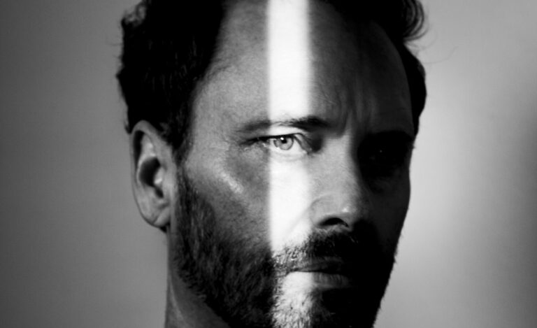 Zwart-wit foto van een man met een intense blik, een kort geschoren baard en een schaduw die zijn gezicht diagonaal verdeelt.