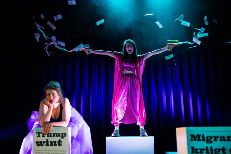 Twee performers op een podium, met een vrouw in een felroze outfit die met twee armen geld of papier in de lucht gooit, terwijl een andere vrouw op een kist zit met teksten op posters.