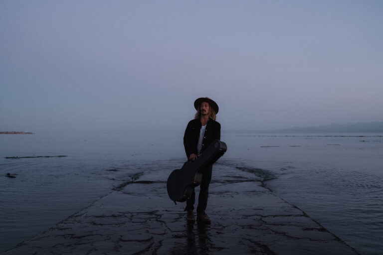 Een persoon met een gitaar staat op een mistige pier bij het water.