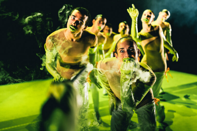 Dansers staan in verschillende poses in gifgroene outfits en lippen op een even groen podium