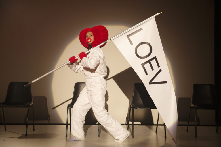 Een performer in een wit kostuum met een rode pruik en gezichtsmake-up, die een vlag met het woord 