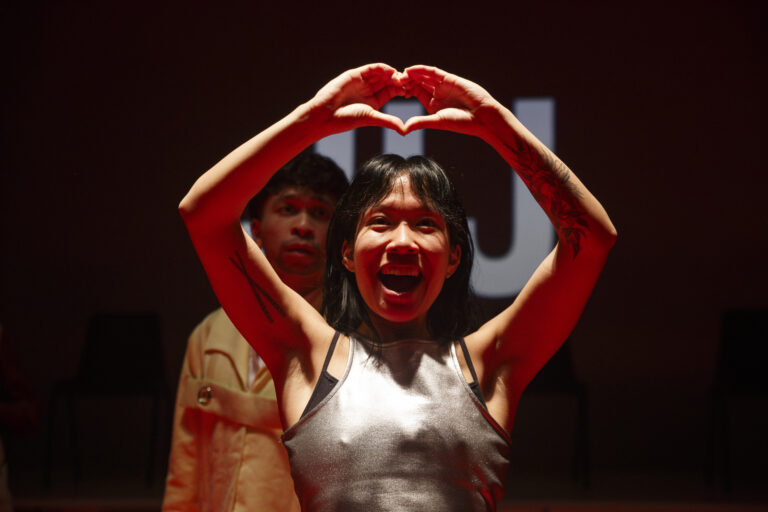 Twee mensen vormen samen een hartvorm met hun armen boven hun hoofden tegen een donkere achtergrond.