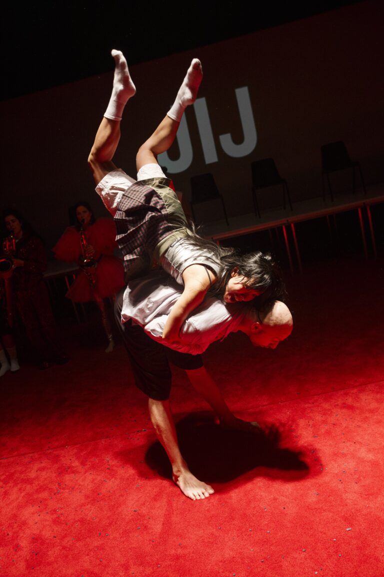 Twee dansers voeren een spectaculaire lift uit op een rode dansvloer tijdens een voorstelling.