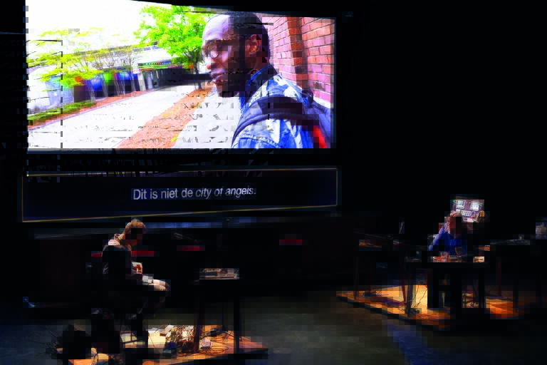Op het podium zitten twee mensen en. Ze kijken naar een projectie van een man