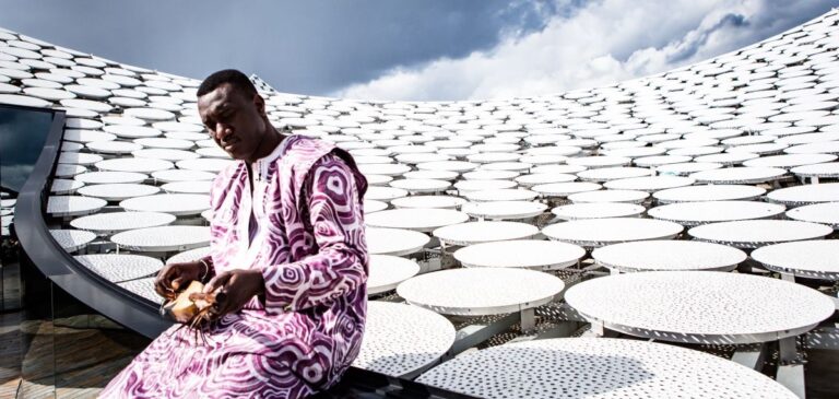Een man in een paars-wit traditioneel Afrikaans gewaad bespeelt een muziekinstrument, zittend op een modern gebouw met een dak bedekt met ronde, witte schijven, onder een bewolkte hemel.