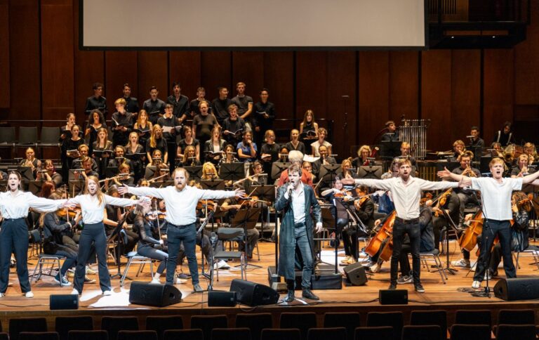 Op de voorgrond de frontzanger gekleed in lange zwarte leren jas, met links en rechts naast hem in wit geklede zangers en zangeressen, die met armen gespreid staan. Achter hen het orkest, daar weer achter een ongeveer 25-koppig koor