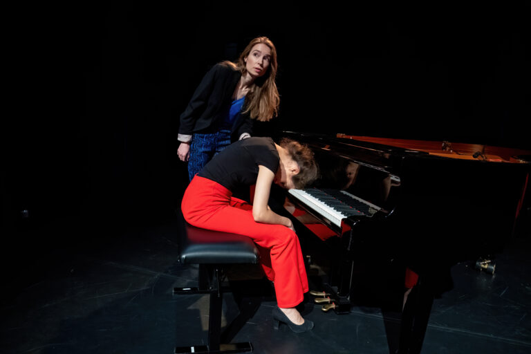 Twee personen bij een vleugelpiano op een donker podium, waarbij één zittend in een rode broek is en de ander naast de piano staat.