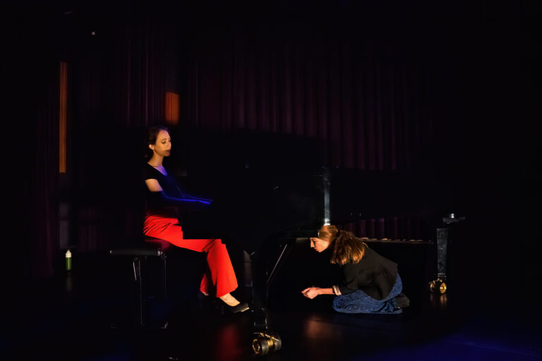 Op een donker podium zit een persoon aan een piano, terwijl een tweede persoon voor de piano op de grond knielt.