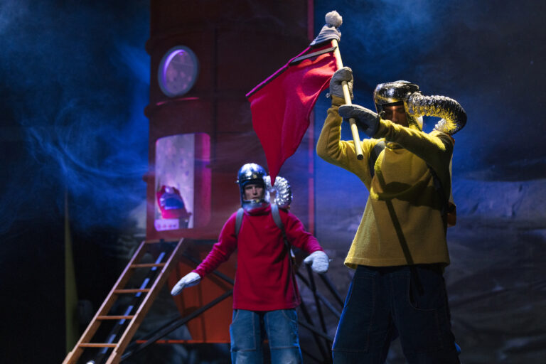 Buurman en Buurman staan in zelfgemaakte ruimtepakken, terwijl één Buurman een vlag vasthoudt en de ander met een serieuze blik bij hun