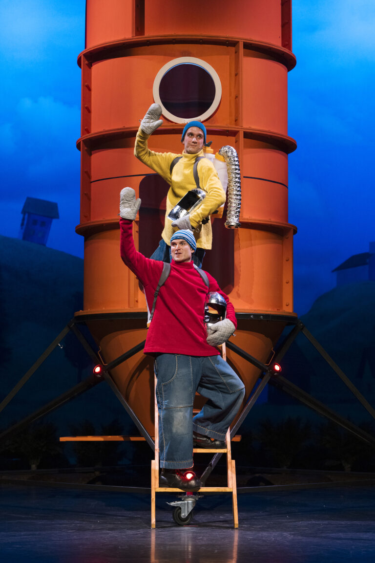 Buurman en Buurman staan in vrolijke kostuums voor een grote oranje raket, poserend met ovenwanten en gereedschap, klaar voor een humoristisch avontuur.