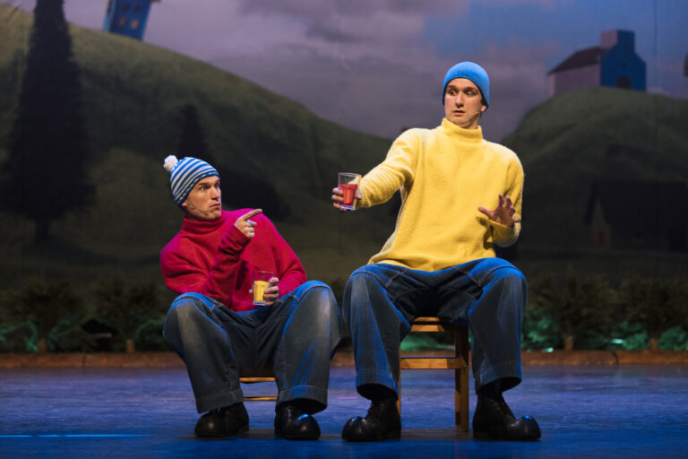 Buurman en Buurman zitten op stoelen met kleurrijke drankjes in hun handen, waarbij één Buurman verbaasd naar het glas van de ander wijst, klaar voor een komische situatie.