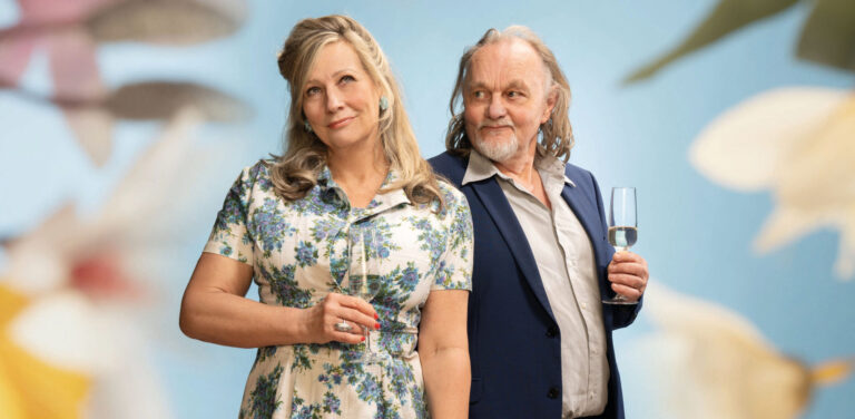 Twee oudere acteurs met glas champagne met een bloemen achtergrond