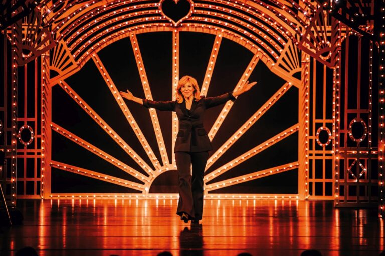 Een performer staat op een verlicht podium onder een grote oranje boog van lampjes met stralende lijnen op de achtergrond.