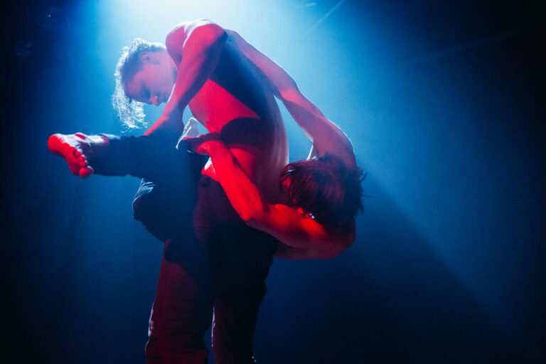 Twee dansers in rode kleding voeren een dansbeweging uit onder blauw podiumlicht.