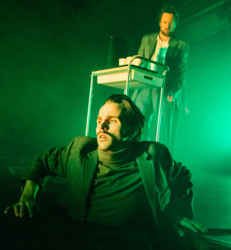Een man in donkere kleding ligt op de vloer van een groen verlicht podium.