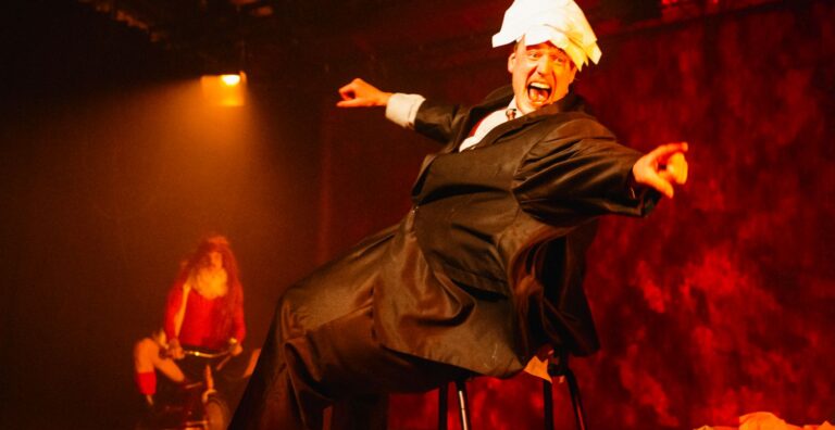 Een performer in smoking en witte pruik voert een dramatische pose uit op een podium tijdens een voorstelling.