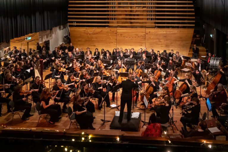 Een grote symfonieorkest speelt in een concertzaal onder leiding van een dirigent.