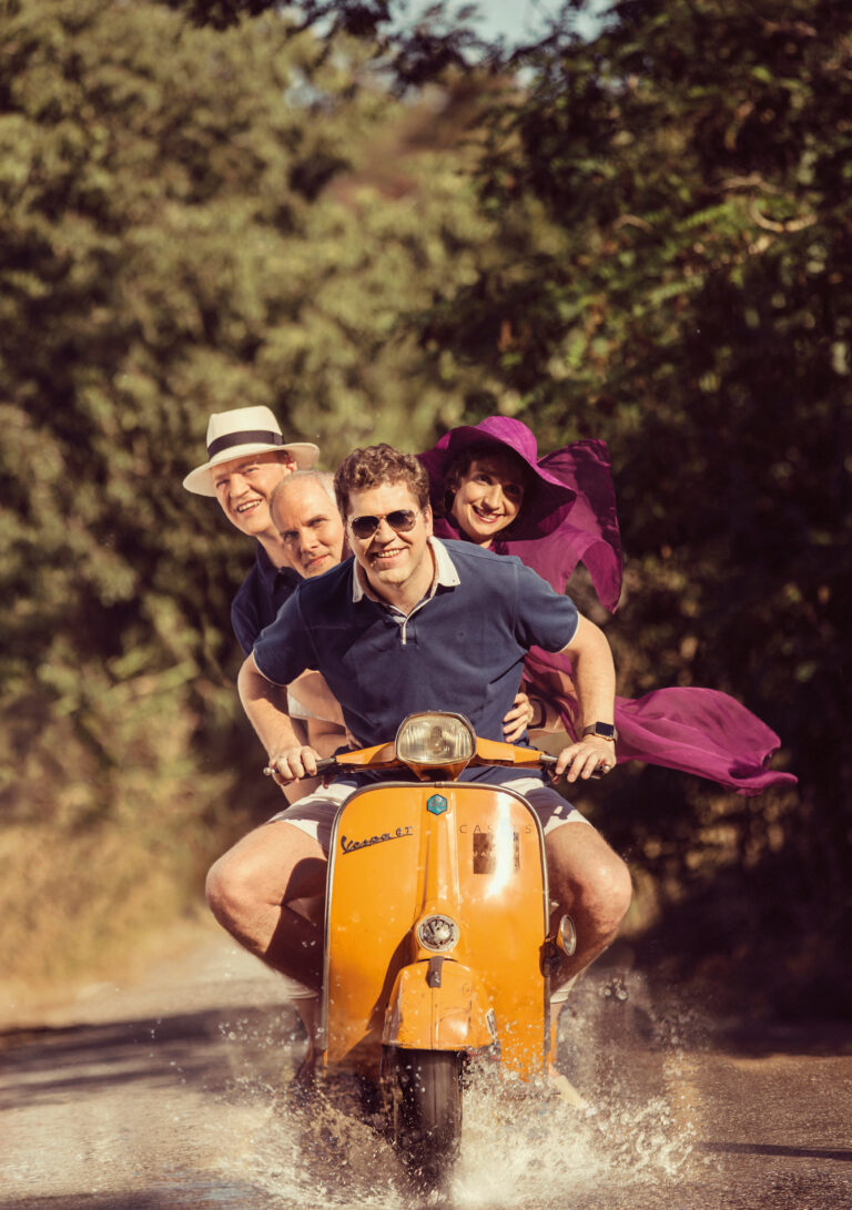Vier muzikanten op een gele scooter rijdens door een plas.