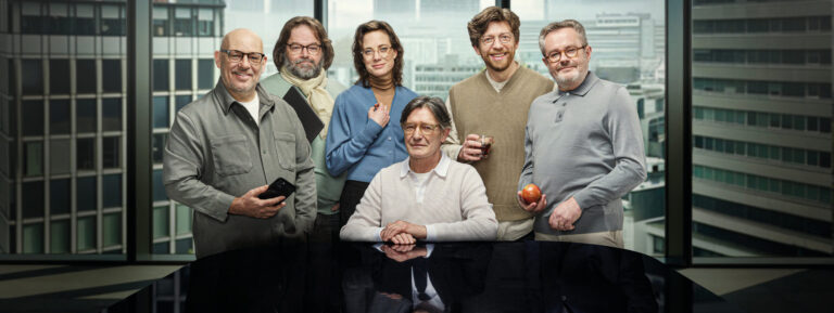 Groepsfoto van alle acteurs in nerdige kleding met een bril op achter een glanzende tafel met acteurs in pak als weerschijning.