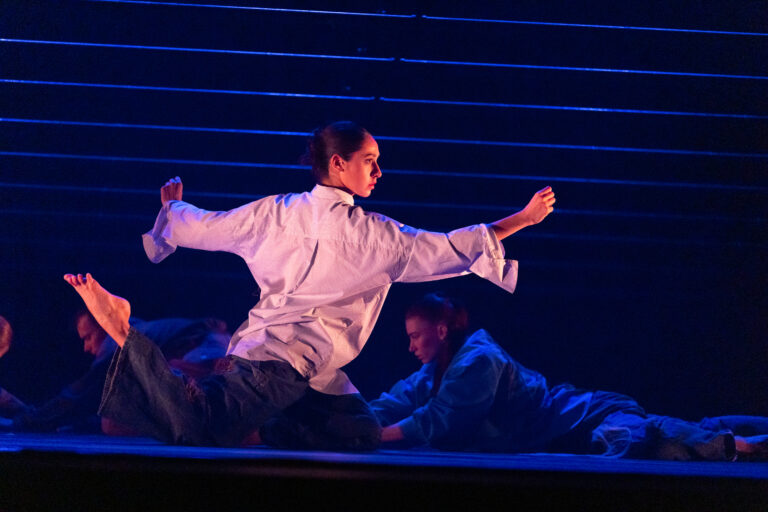 Een danser in een wit shirt voert een uitgestrekte, dynamische pose uit op een donker podium met blauwe verlichting.
