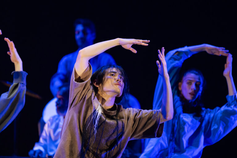 Een danser op een podium met één arm omhoog, omgeven door blauw podiumlicht.
