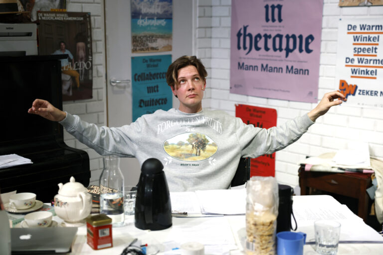 Acteur zit achter een rommelig bureau met zijn handen omhoog