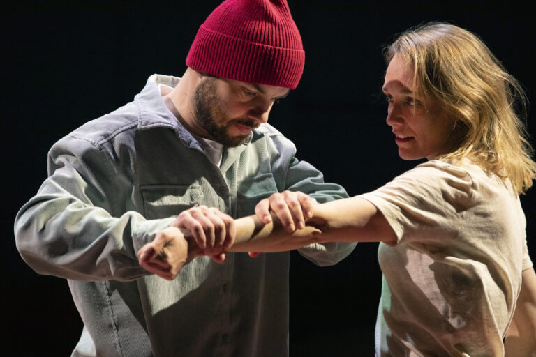 Een man met een rode muts en een vrouw in een licht shirt zijn bezig met een intieme interactie op het podium.