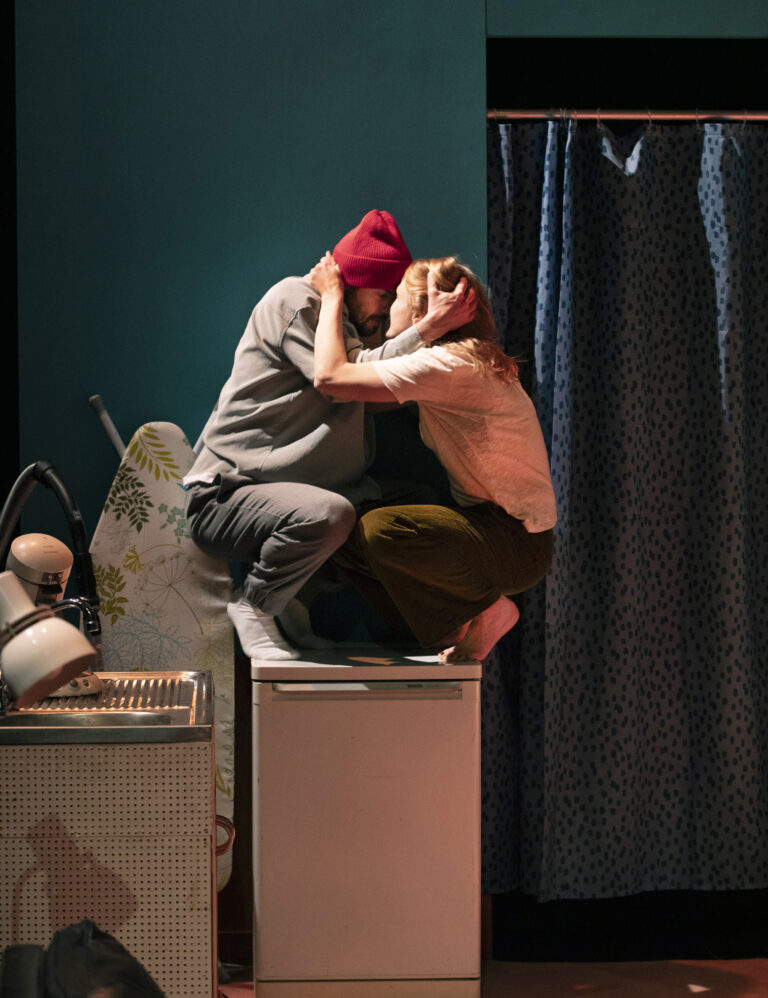 Een man en een vrouw omarmen elkaar terwijl ze op een keukenoppervlak zitten in een opstelling met een blauwe muur en een gordijn op de achtergrond.