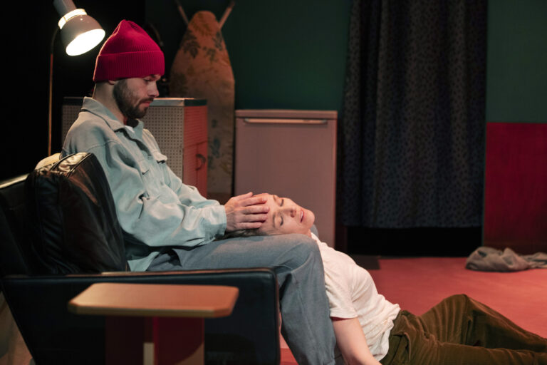 Een man in een rode beanie zit op een zwarte stoel en houdt de hoofden van een tweede persoon, die op de grond ligt, terwijl ze in een uitdagende ruimte zijn.