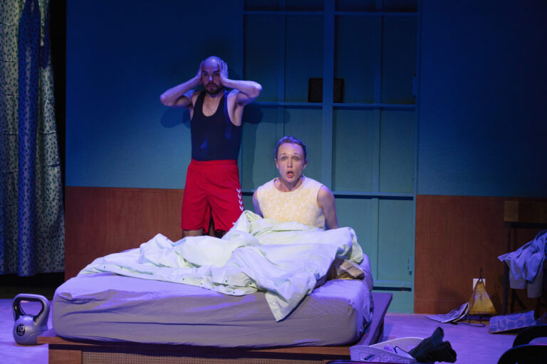 Twee mannen op een bed in een blauw verlichte kamer; één staat naast het bed en de ander zit onder een dekbed.