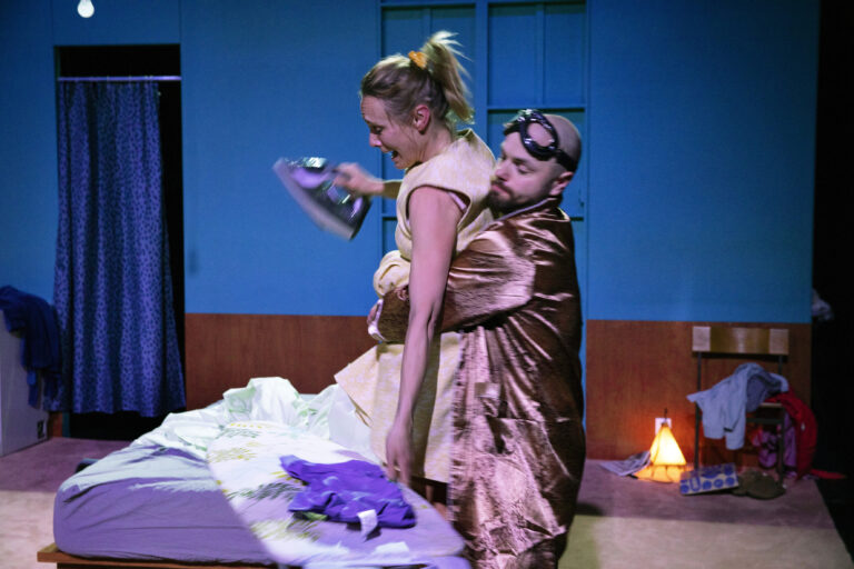 Twee vrouwen staan bij een bed in een slaapkamerachtige scène op een podium, waarbij een vrouw een satijnen roze-bruin jurk draagt.