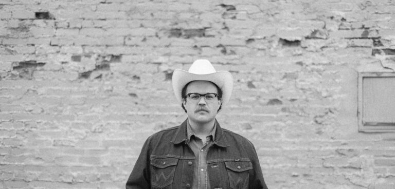 Een man met een snor en een bril staat voor een verweerde bakstenen muur, gekleed in een spijkerjas en -broek, met een grote riemgesp en een witte cowboyhoed.